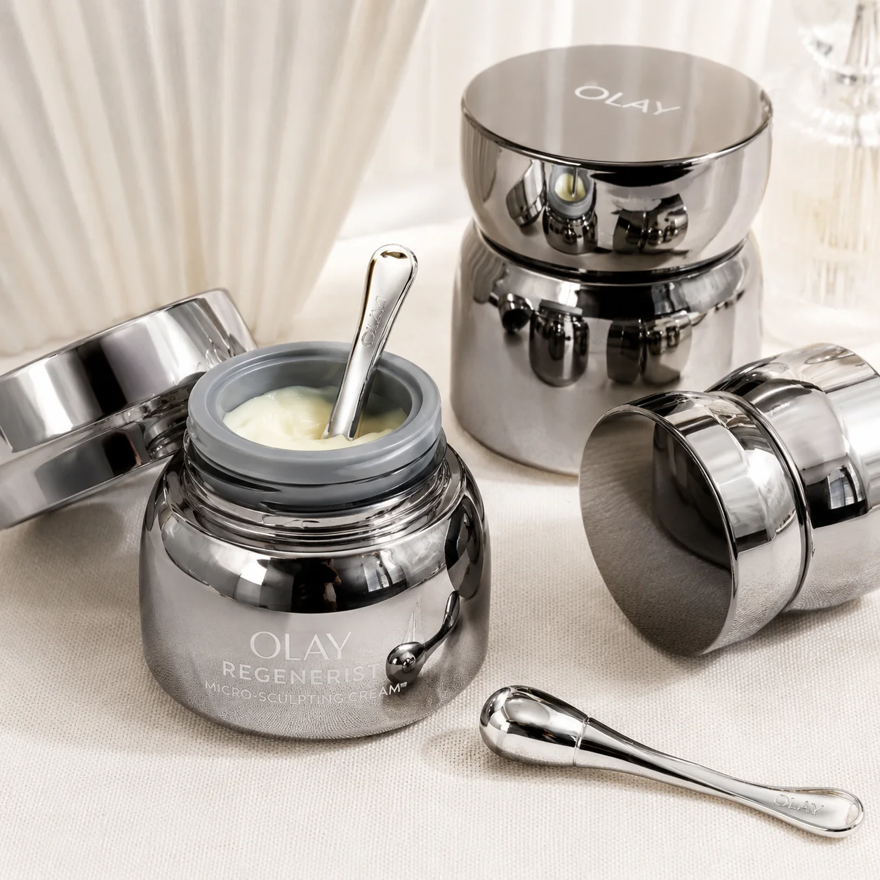 Olay Regenerist Micro-Sculpting Cream in sleek chrome jars, with one jar open, revealing cream and a metal applicator on a cream fabric background.